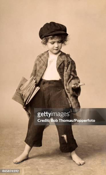 Posed studio portrait of a raggedy young barefoot child selling newspapers in London, circa 1920.