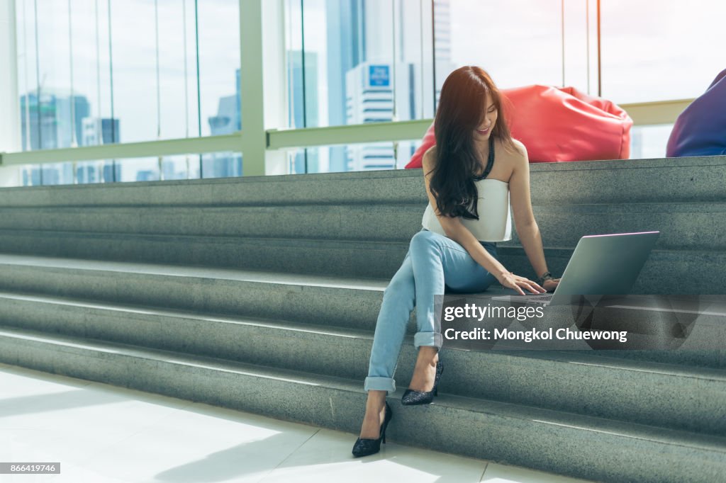 Beautiful young asian woman is using a laptop and smiling while sitting on stairs