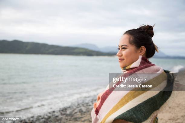 māori woman wrapped in a shawl looks out to sea on a beautiful beach - woman in a shawl stockfoto's en -beelden