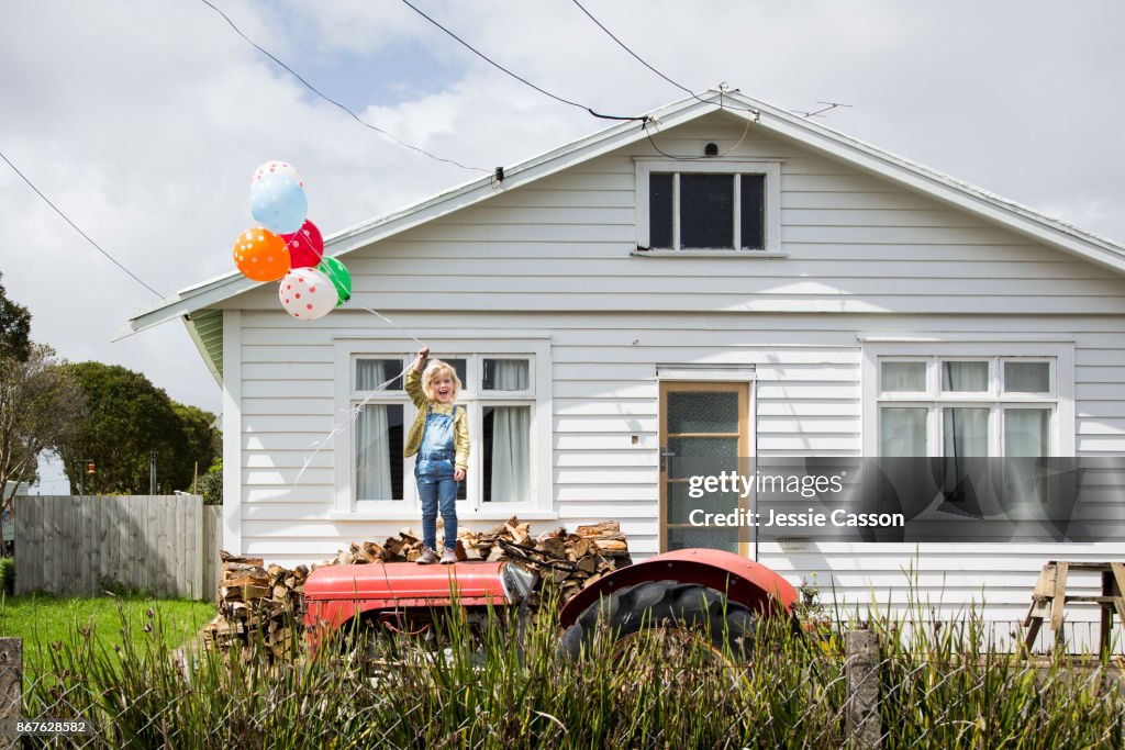 Girl stands on a tractor holding colourful balloons in front of a bungalow