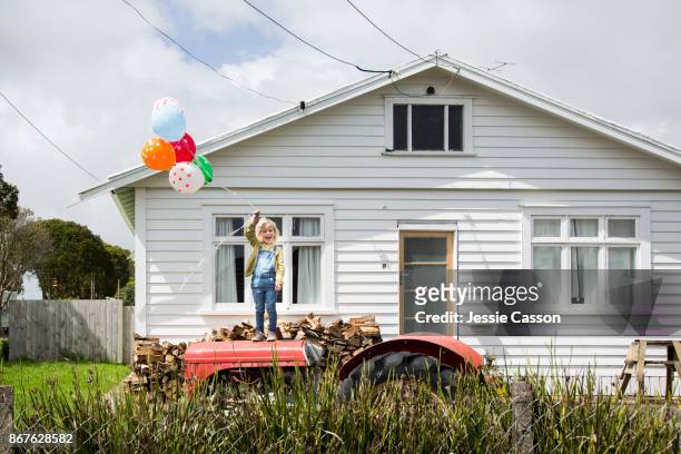 girl stands on a tractor holding colourful balloons in front of a bungalow - new zealand housing stock pictures, royalty-free photos & images