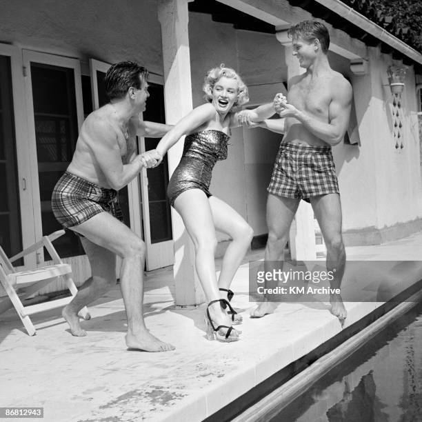 American actress Marilyn Monroe wearing heels and a bathing suit, plays around with two male friends at the pool, Hollywood, California, 1950.