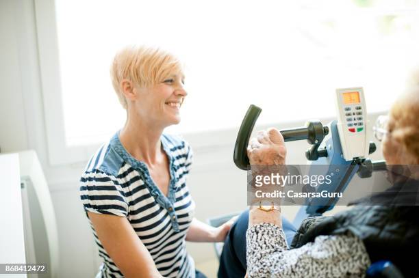 therapist helping senior woman on wheelchair doing exercises on exercising bike - active retirement community stock pictures, royalty-free photos & images