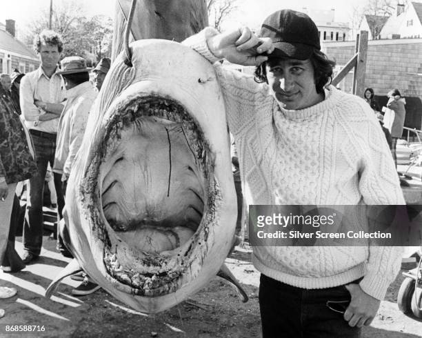 Portrait of American film director Steven Spielberg as he poses beside a shark on a hook, mid 1970s.