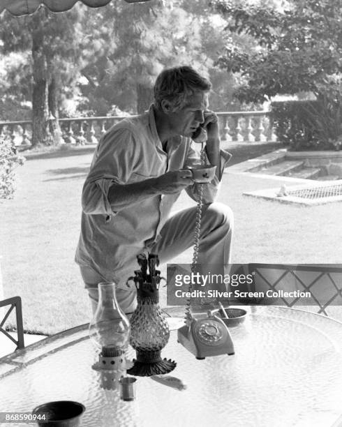 American actor Steve McQueen holds a cup as he talks on the telephone at an outdoor table in his garden, Brentwood, Los Angeles, California, 1960s.