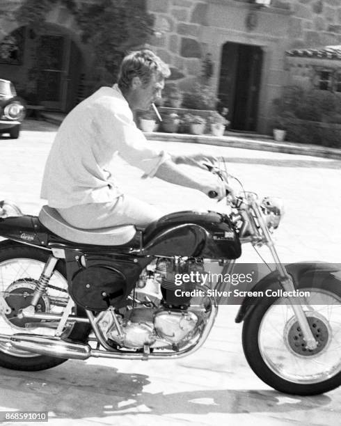 View of American actor Steve McQueen , a cigarette in his mouth, as he sits on a motorcycle in his driveway, Brentwood, Los Angeles, California,...