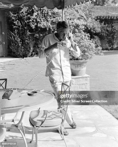 American actor Steve McQueen holds a cup as he talks on the telephone at an outdoor table in his garden, Brentwood, Los Angeles, California, 1960s.