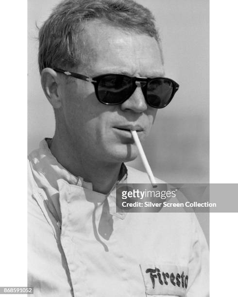 View of American actor Steve McQueen , in Firestone racing suit and sunglasses, as he smokes a cigarette at Riverside Raceway, Riverside, California,...