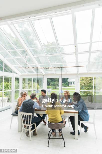 multi raciale groupe d’amis assis autour de la table dans la salle de soleil profiter de dîner - conservatory house photos et images de collection