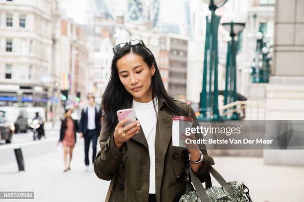 asian businesswoman looks at smart phone while walking in city. - coffee bag stock-fotos und bilder