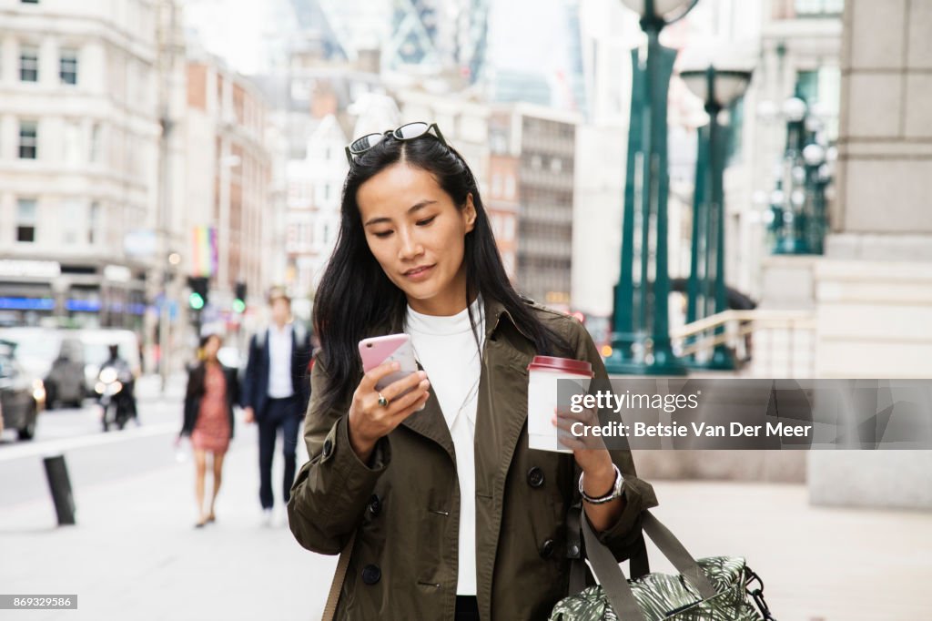 Asian businesswoman looks at smart phone while walking in city.