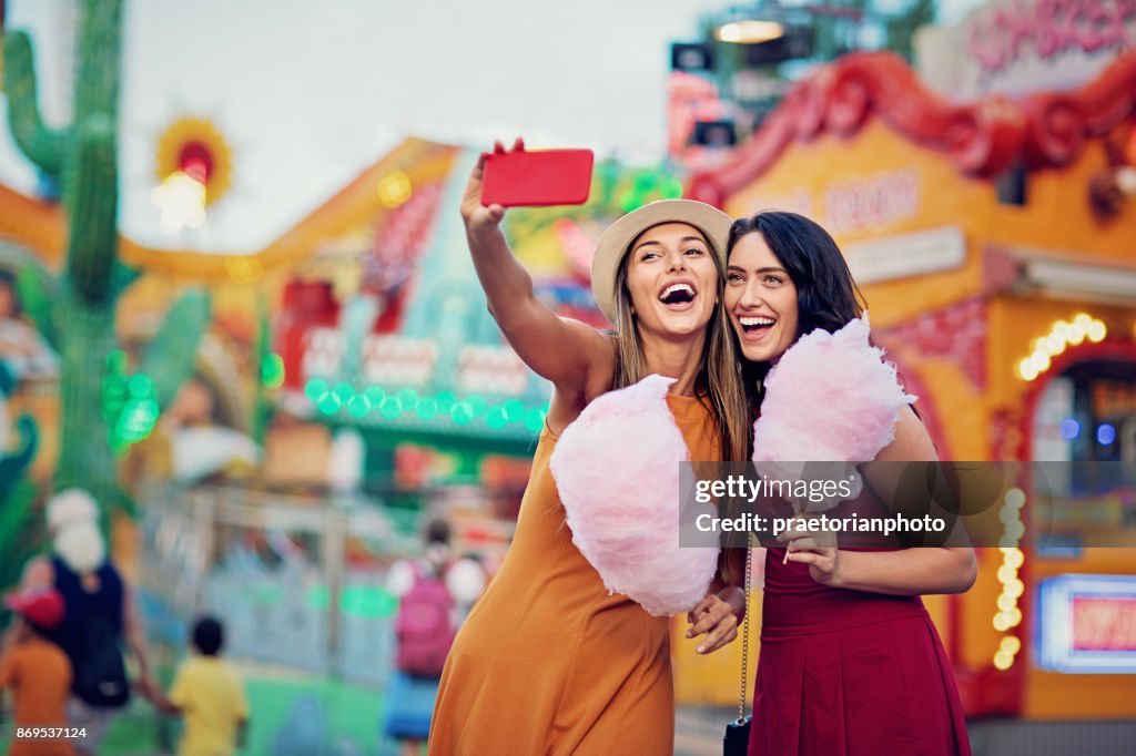 Happy girlfriends are taking selfie/making video call and make fun together in fun fair
