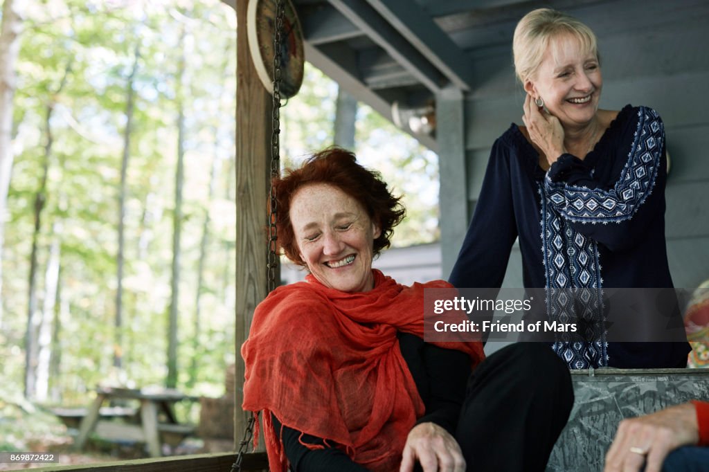 Two senior women sit on cabin porch in woods and laugh