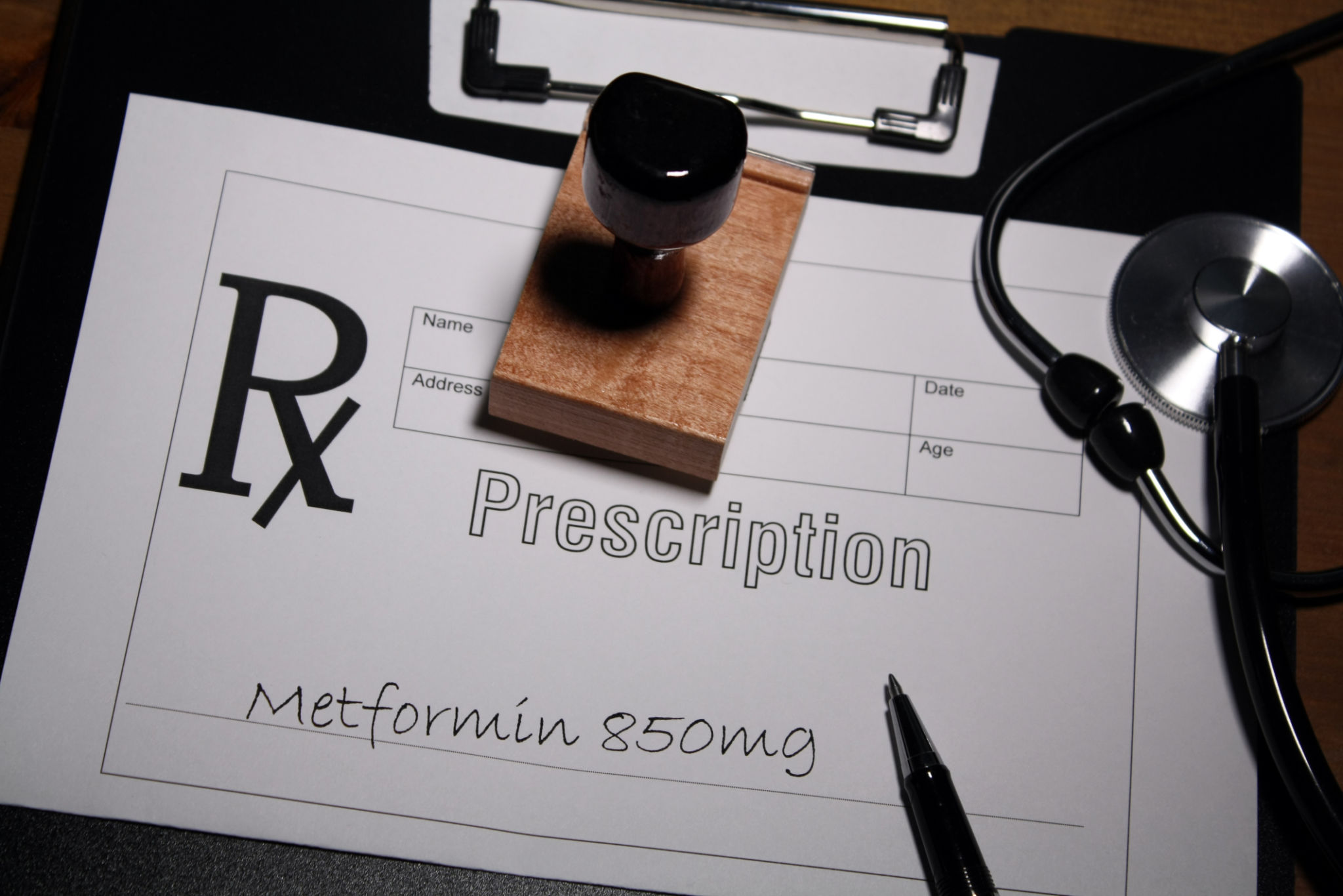 A medical prescription form labeled 'Prescription' with 'Metformin 850mg' handwritten on it, alongside a wooden stamp, a stethoscope, a pen, and a clipboard. A medical prescription form labeled 'Prescription' with 'Metformin 850mg' handwritten on it, alongside a wooden stamp, a stethoscope, a pen, and a clipboard.