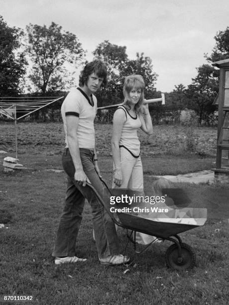 English footballer Mike Channon and his wife Jane push their baby daughter Nicola in a wheelbarrow while working on their farm, UK, August 1973.
