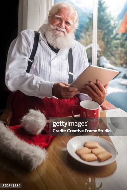 happy santa claus holding a digital tablet while having an afternoon tea with cookies  it's cold and snow out site - happy santa claus over white blank stockfoto's en -beelden