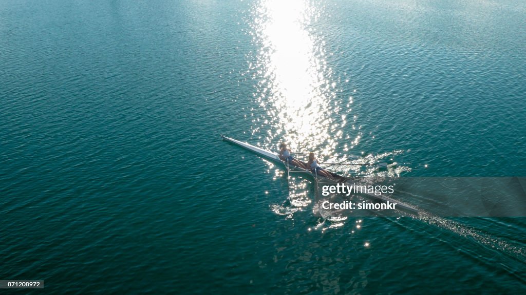 Two female athletes sculling on lake in sunshine