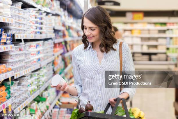woman reads label on yogurt while grocery shopping - etiqueta nutricional imagens e fotografias de stock