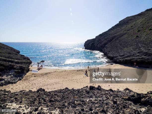 people bathing and sunbathing on the beach. rocky coast of the cabo de gata with formations of volcanic rock of black color and beach of sand of brown color. cabo de gata - nijar natural park, cala chica, almeria, andalusia, spain. - cabo de gata fotografías e imágenes de stock