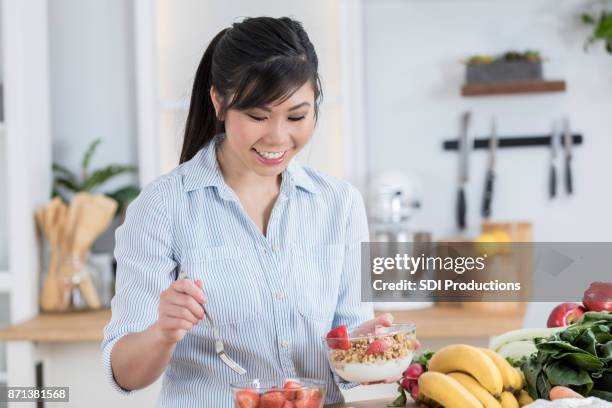 cheerful young woman adds fruit to a yogurt parfait - fruit parfait stock pictures, royalty-free photos & images