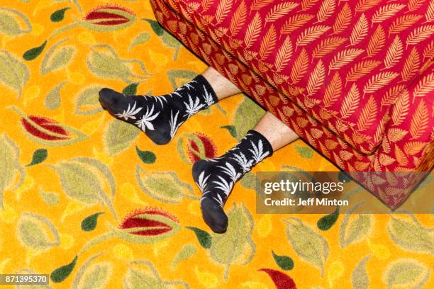 humorous view of a persons feet sticking out from under a brightly colored and pattered sofa while laying on a brightly colored leaf print rug - off beat stock pictures, royalty-free photos & images