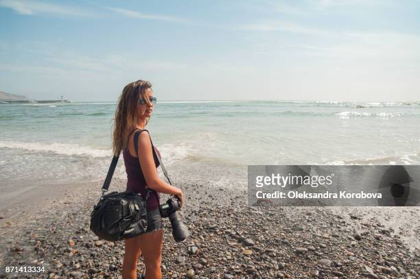 young woman photographer enjoying peaceful seaside vacation, standing on the beach by the ocean on a sunny day. - lima stockfoto's en -beelden