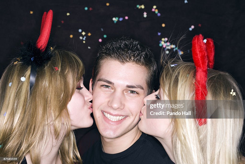 Girls wearing devils' horns kissing a boy, smiling