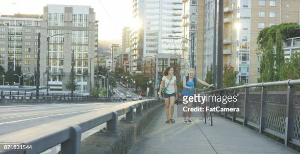 zwei attraktive junge frauen fuß fahrrad auf bürgersteig an einem sonnigen nachmittag - ameisenstraße stock-fotos und bilder