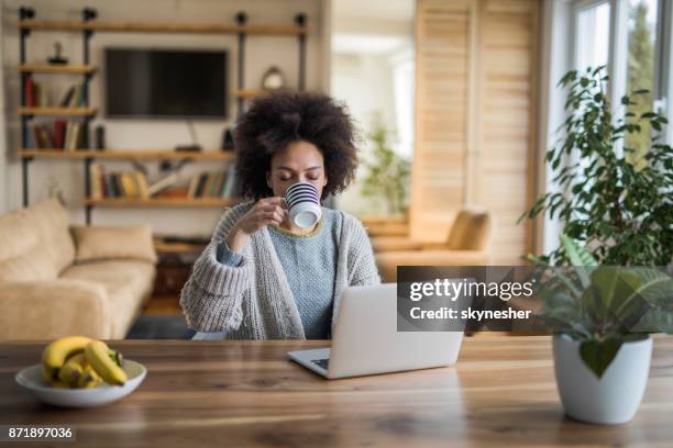 young african american woman drinking coffee and using laptop at home. - black mug stock pictures, royalty-free photos & images