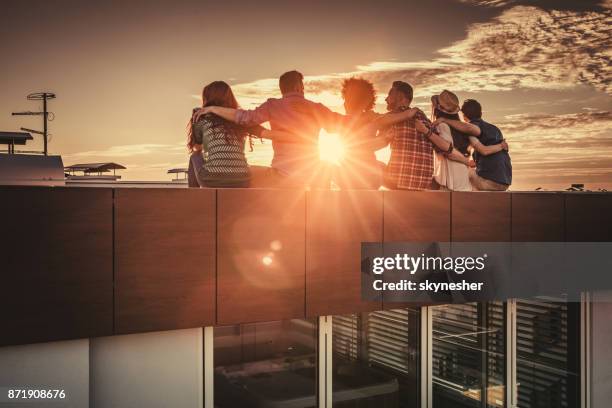 rear view of embraced friends relaxing on a rooftop at sunset. - friends sunset imagens e fotografias de stock