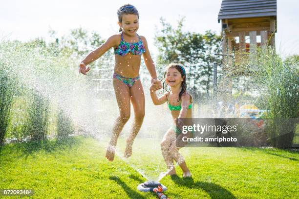 jump - happy young sisters playing in garden jumping over sprinkler on summer afternoon - jumping sprinkler stock pictures, royalty-free photos & images