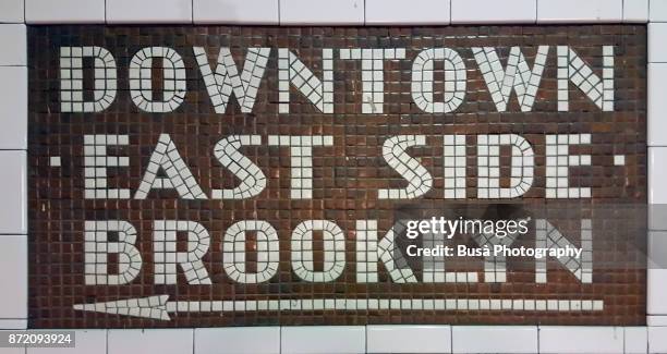 new york city, ny, usa - october 10, 2017: colorful ceramic plaques and tile mosaics in the new york city subway. mosaic directing passengers in direction downtown - east side - brooklyn - underground sign 個照片及圖片檔