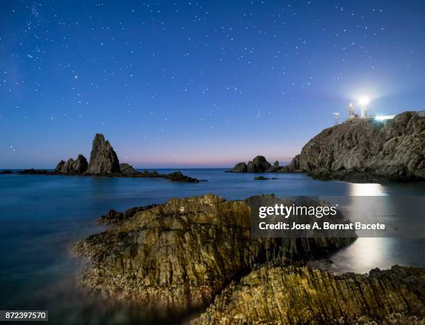 sea landscape at night, with reef rocks in the water and a lighthouse illuminating the beach, a night of stars with blue sky. cabo de gata - nijar natural park, sirens reef,  biosphere reserve, almeria,  andalusia, spain - lighthouse reef - fotografias e filmes do acervo