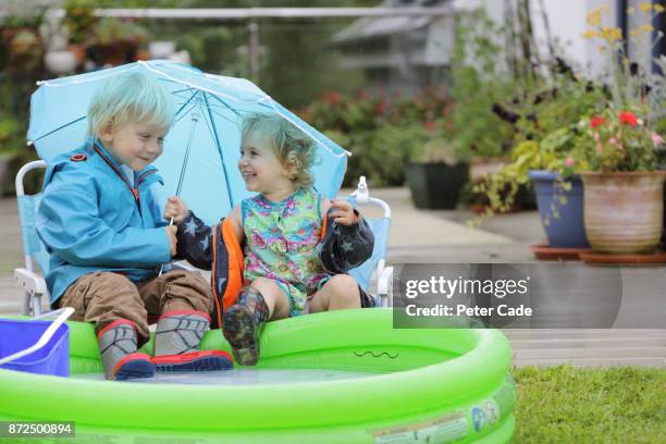 young boy and girl sitting next to paddling pool outside in the rain under umbrella - brother sister shower stock-fotos und bilder