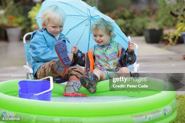 young boy and girl sitting next to paddling pool outside in the rain under umbrella - water repellent stock pictures, royalty-free photos & images