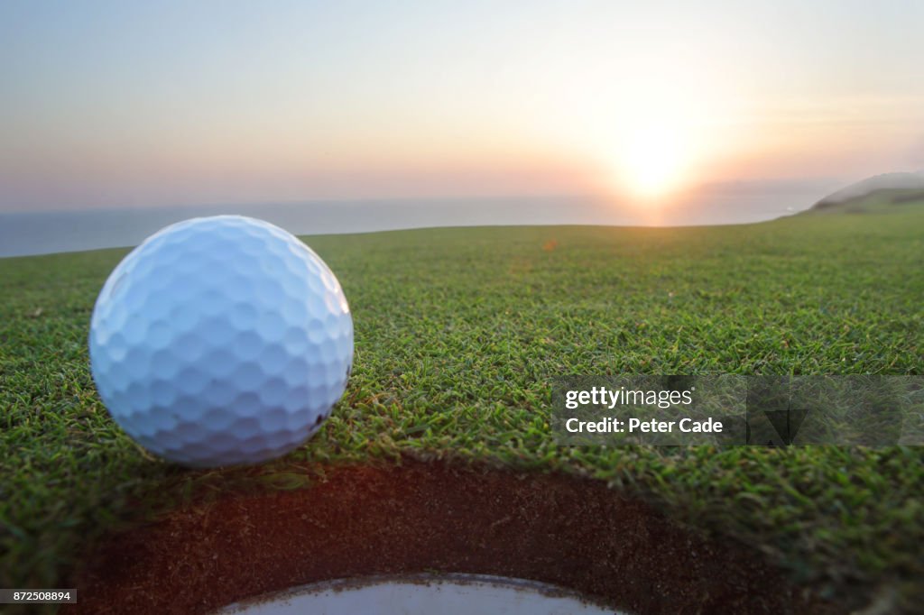 Golf ball sitting on edge of hole at sunset