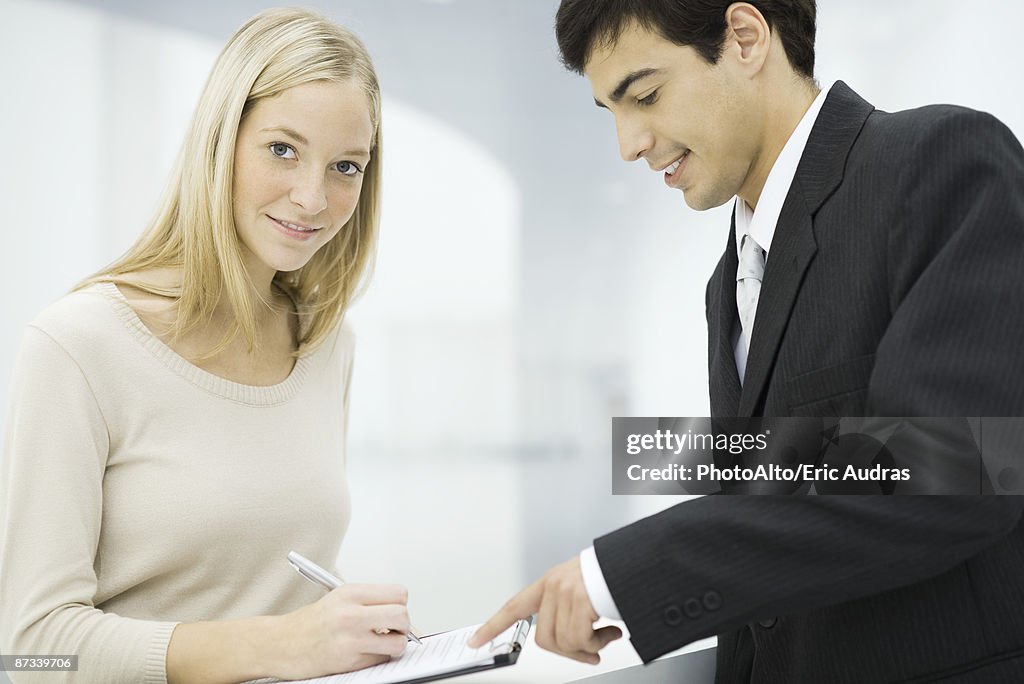 Businessman holding out clipboard, woman writing on document and smiling at camera