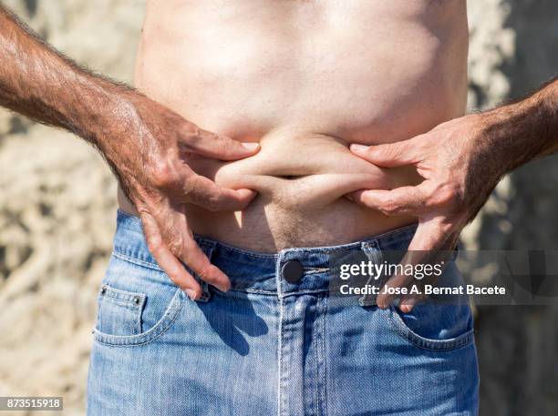 a close-up image of a men mature with  belly fat pinching itself, white-skinned, with jeans shorts in the light of the sun in the beach - pot belly fotografías e imágenes de stock