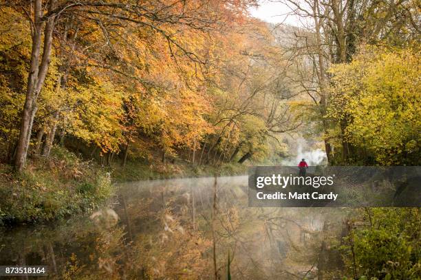 Man on his bike passes the autumn colours from the trees reflected in the water of the Kennet and Avon canal near the Dundas Aqueduct as the sun...