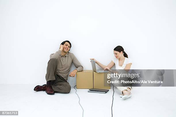 man talking on phone, sitting on floor next to woman using laptop computer at cardboard box desk - makeshift desk stock pictures, royalty-free photos & images