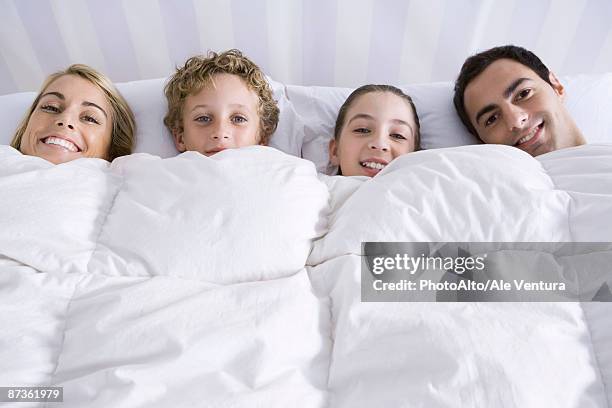 family lying together in bed beneath comforter, smiling at camera - covers head with pillow stock-fotos und bilder