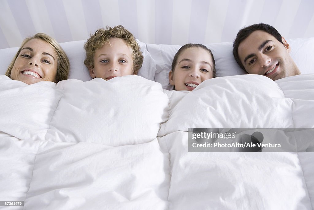 Family lying together in bed beneath comforter, smiling at camera