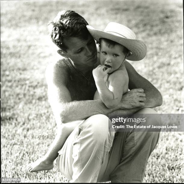 View of lawyer Robert F Kennedy as he holds his son, David , McLean, Virginia, 1958. Both are shirtless; David wears a straw hat.