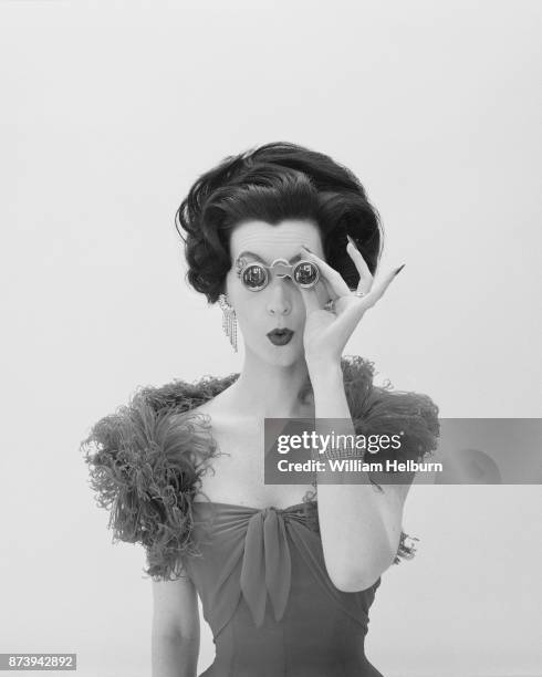 American model Dovima , in a gown with feathered trim, looks through a pair of opera glasses as she purses her lips, New York, New York, 1961. The...