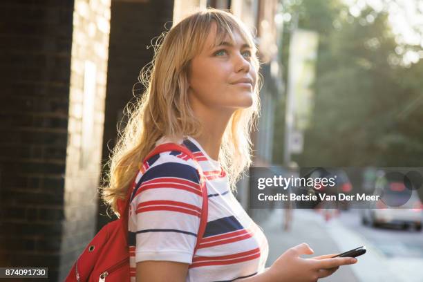 woman looks up from smart phone, standing in city street. - blond hair young woman sunshine stockfoto's en -beelden