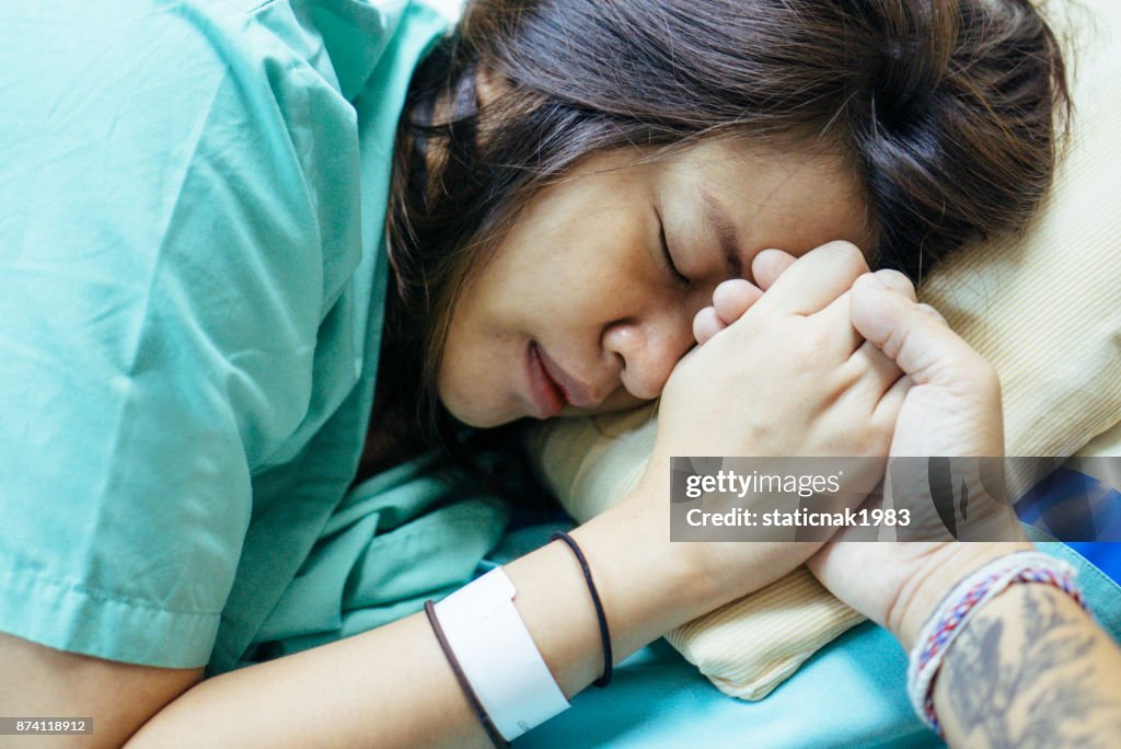 Young man holding hand of asian pregnant woman waiting in hospital bed