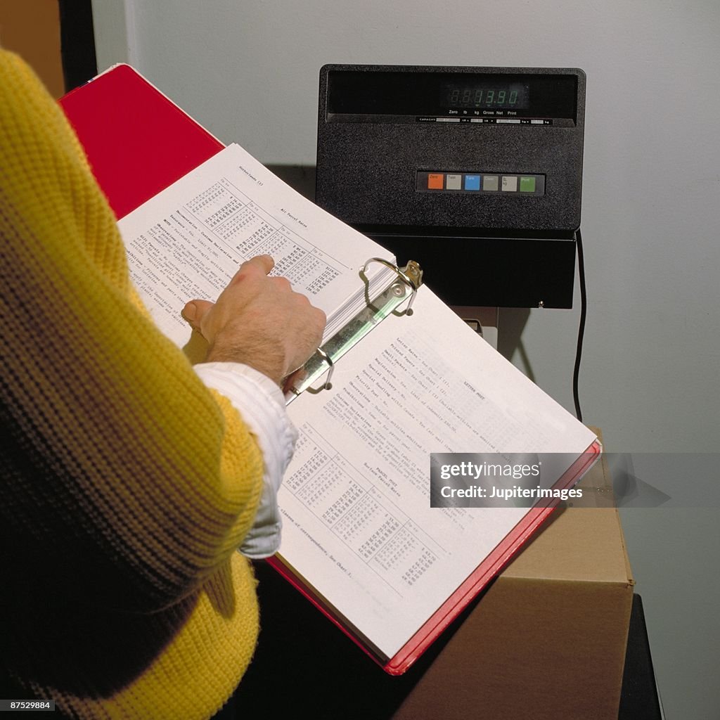 Person with looseleaf notebook weighing parcel for shipping
