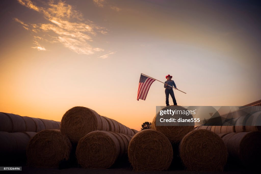 Vaqueira jovem fica no topo de uma enorme pilha de fardos de feno ao pôr do sol, segurando a bandeira americana