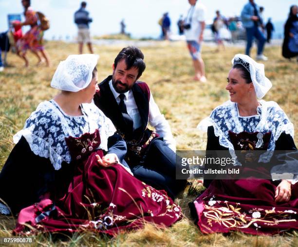 Zwei junge Frauen und ein Mann in traditioneller bretonischer Tracht, die nur noch zu festlichen Anlaessen getragen wird, sitzen plaudernd im...