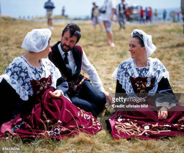Zwei junge Frauen und ein Mann in traditioneller bretonischer Tracht, die nur noch zu festlichen Anlaessen getragen wird, sitzen plaudernd im...
